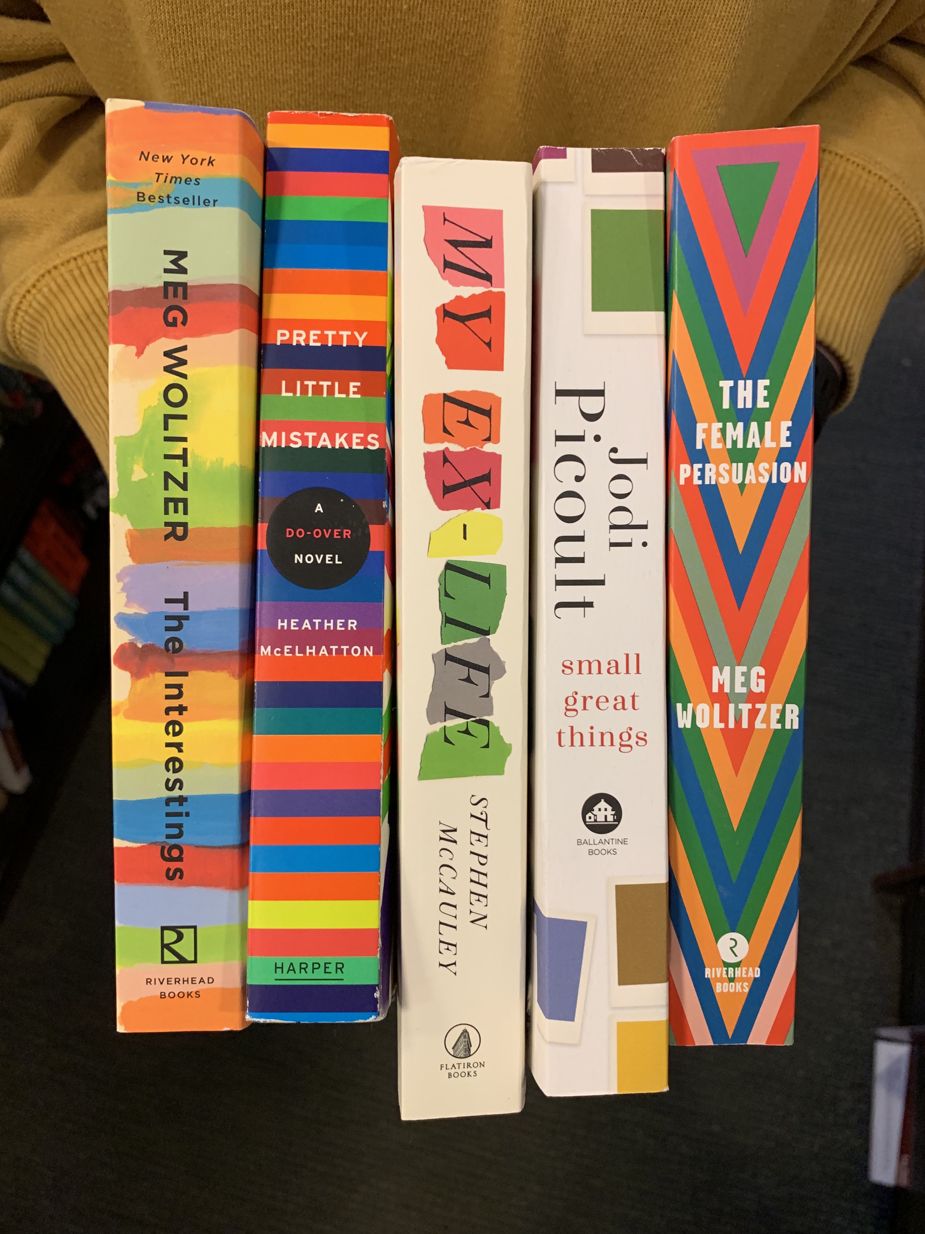 image of me holding five books: "the interestings," "pretty little mistakes," "my ex-life," "small great things," and "the female persuasion" all of them have brightly colored spines